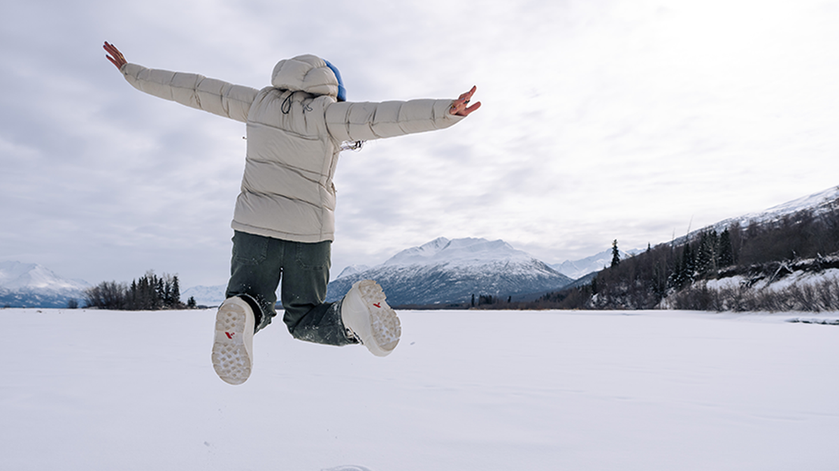 person jumping in winter weather on frozen lake with snowy mountain in background