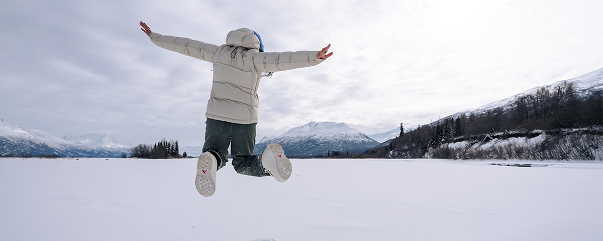 person jumping in winter weather on frozen lake with snowy mountain in background