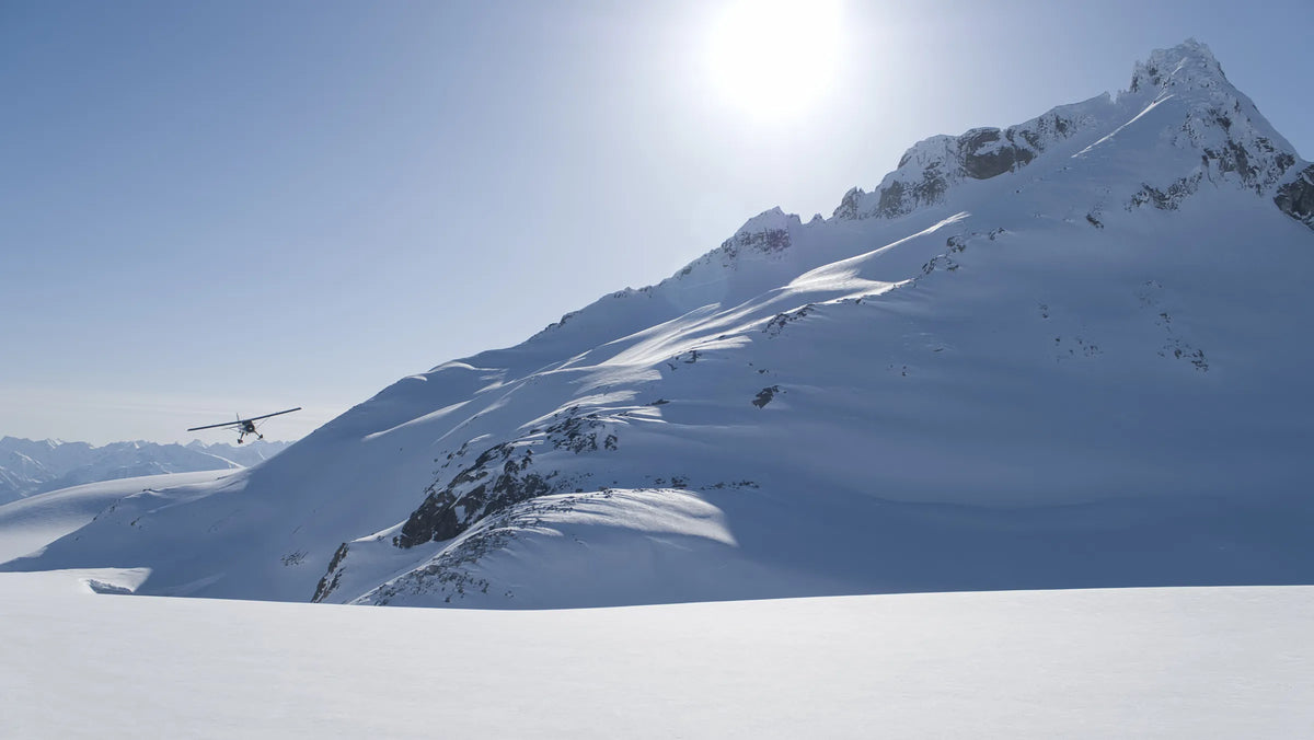A bush plane flies low over a sunlit glacier beside a jagged, snow-covered peak in the Alaska mountains.