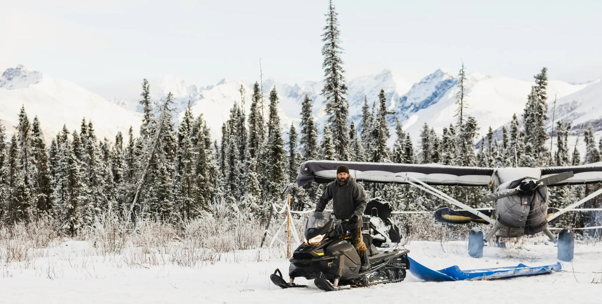 Man in a dark winter jacket, bunny boots and beanie sitting on a black snowmobile towing a blue sled, next to a ski-equipped bush plane with its engine covered, surrounded by frosted spruce trees and snow-covered Alaska mountains. Cold winter background. 